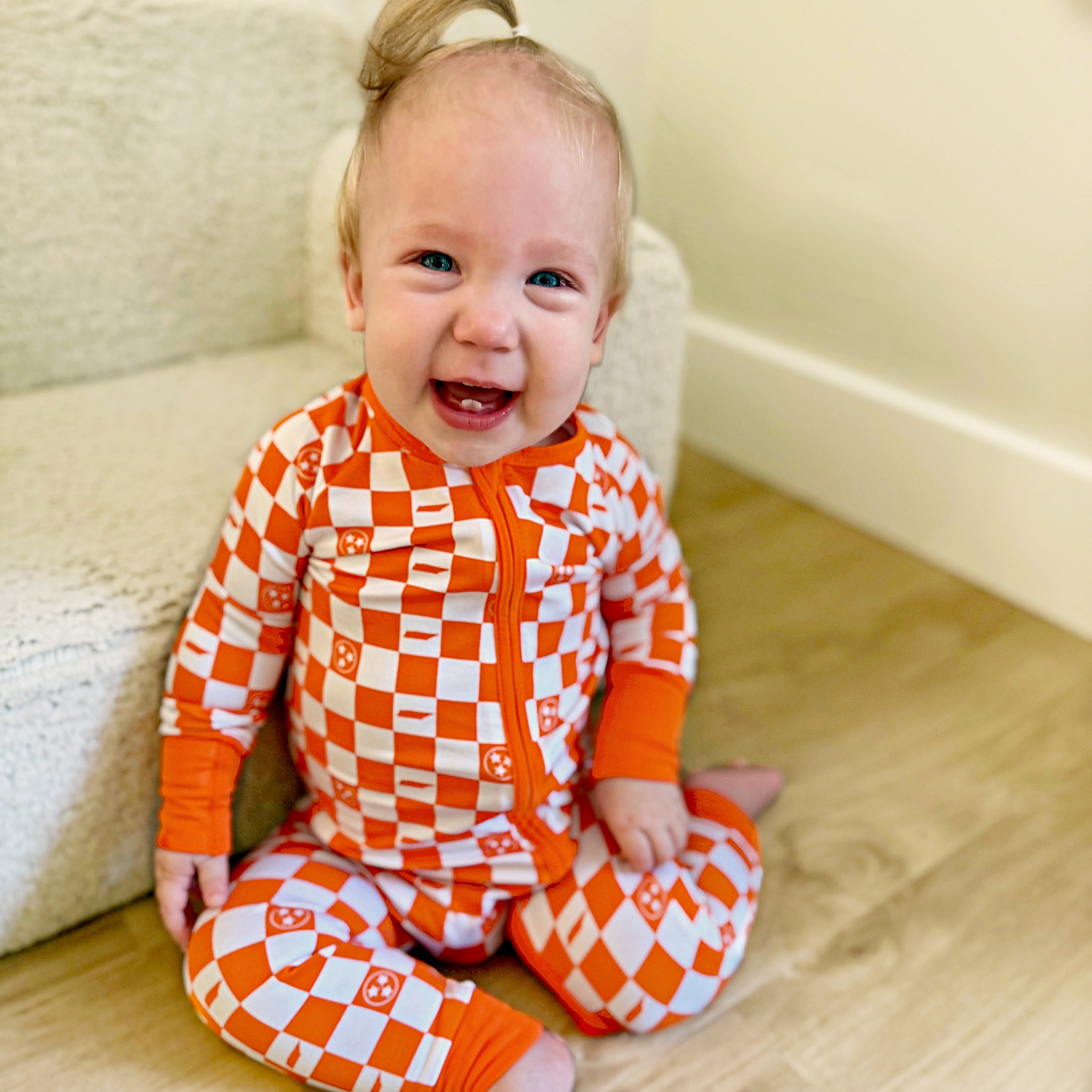 Baby wearing orange and white tennessee checkered pajamas sitting on a wooden floor.