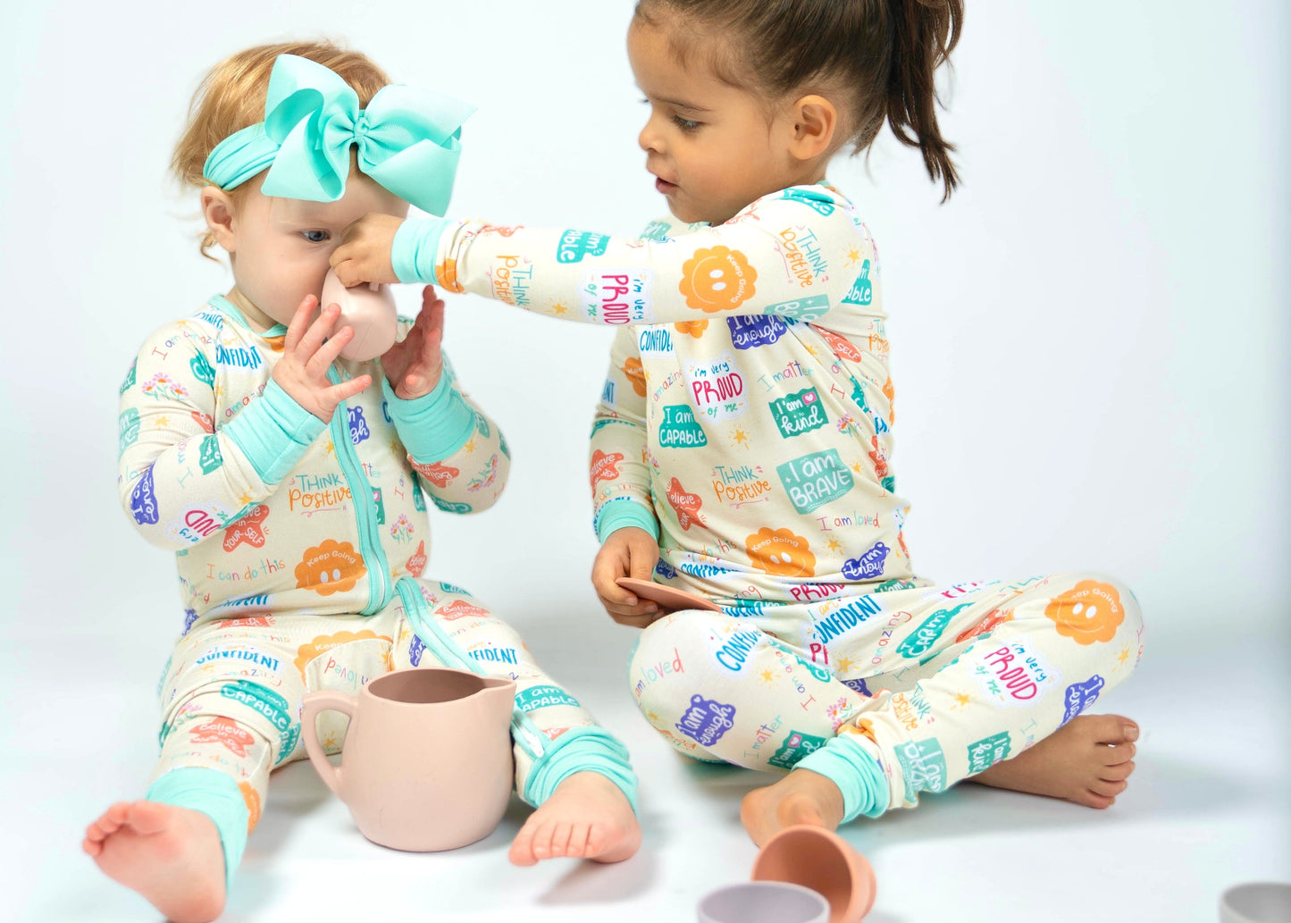 Two children wearing colorful pajamas with positive affirmation designs, sitting on a white floor.