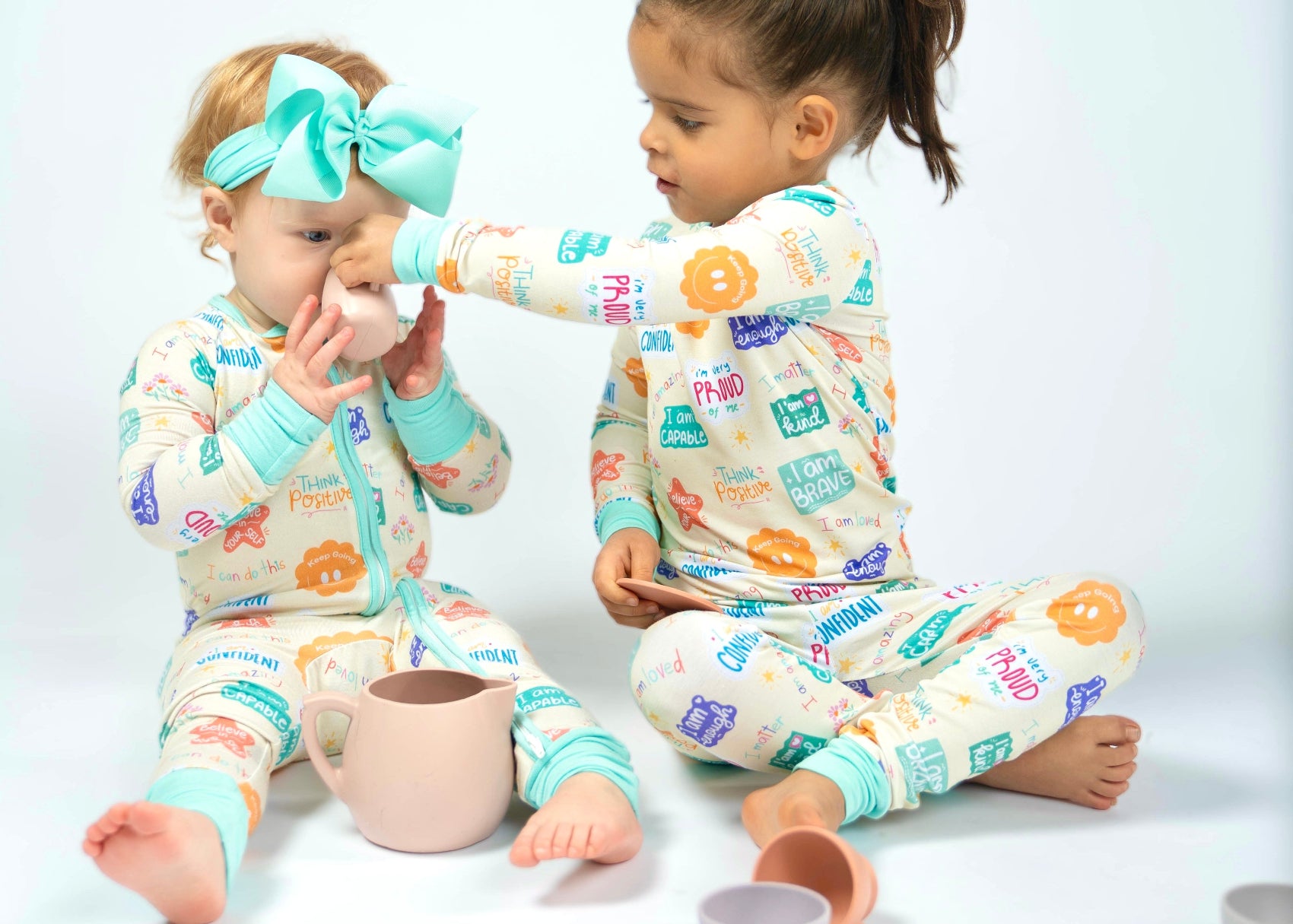 Two children wearing colorful pajamas with positive affirmation designs, sitting on a white floor.
