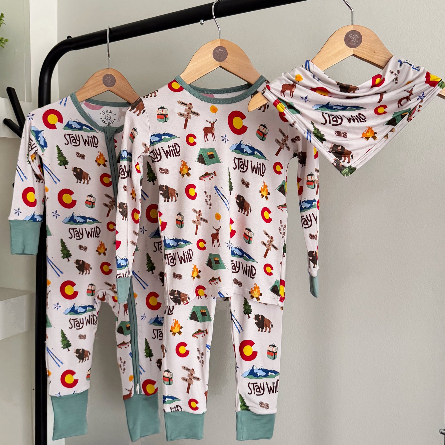 Children's pajamas and bibwith Colorado nature-themed design on hangers against a white wall.