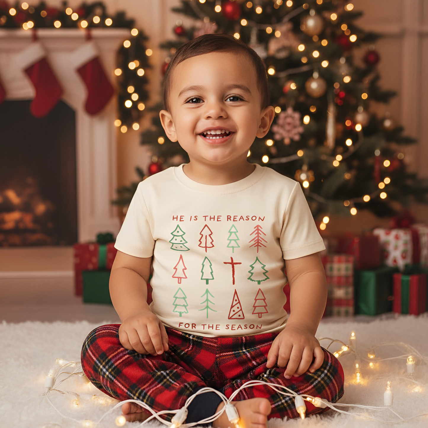 Child wearing a Christmas-themed shirt sitting in front of a decorated Christmas tree and fireplace.