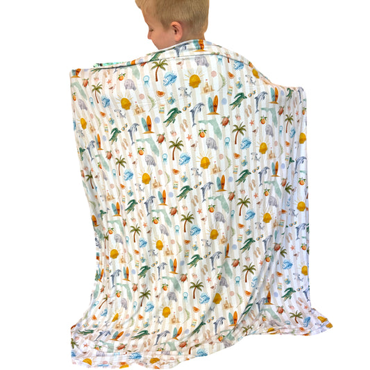 A child wearing a white blanket with a colorful tropical beach print, shown from behind against a plain white background.