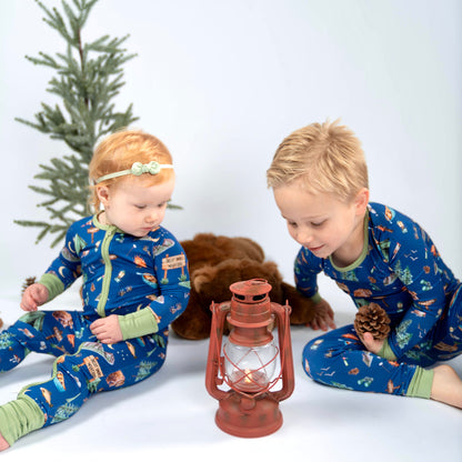 Two children in blue pajamas with nature patterns sitting on a white floor with a small Christmas tree and lantern.