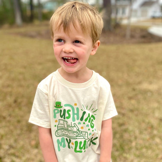 Child wearing a white t-shirt with pushing my luck green text and graphics outdoors