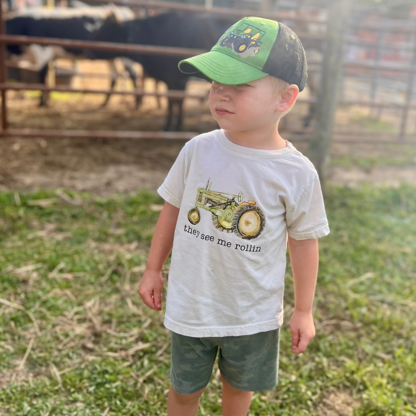 Child wearing a cap and t-shirt with a tractor graphic on a farm.