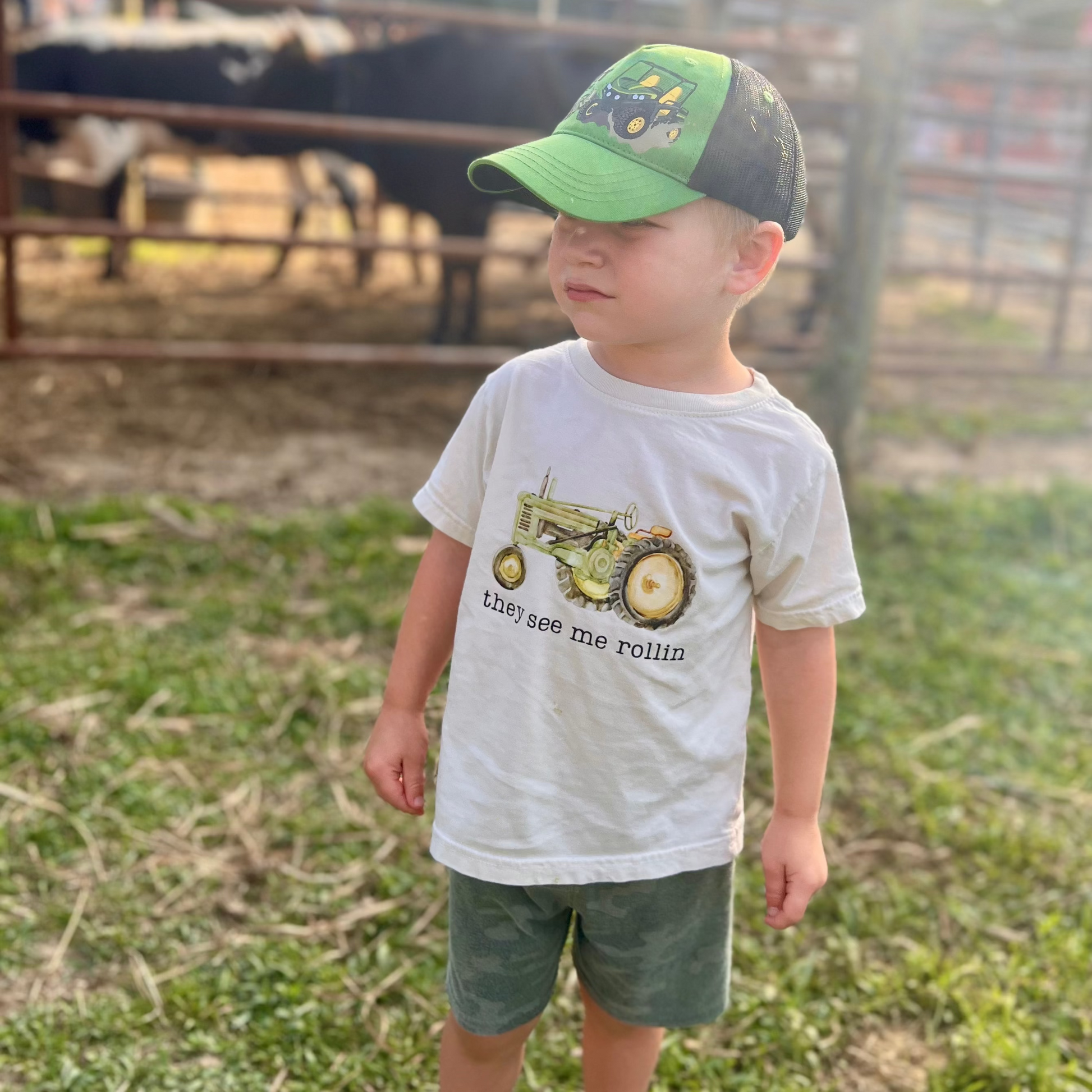 Child wearing a cap and t-shirt with a tractor graphic on a farm.