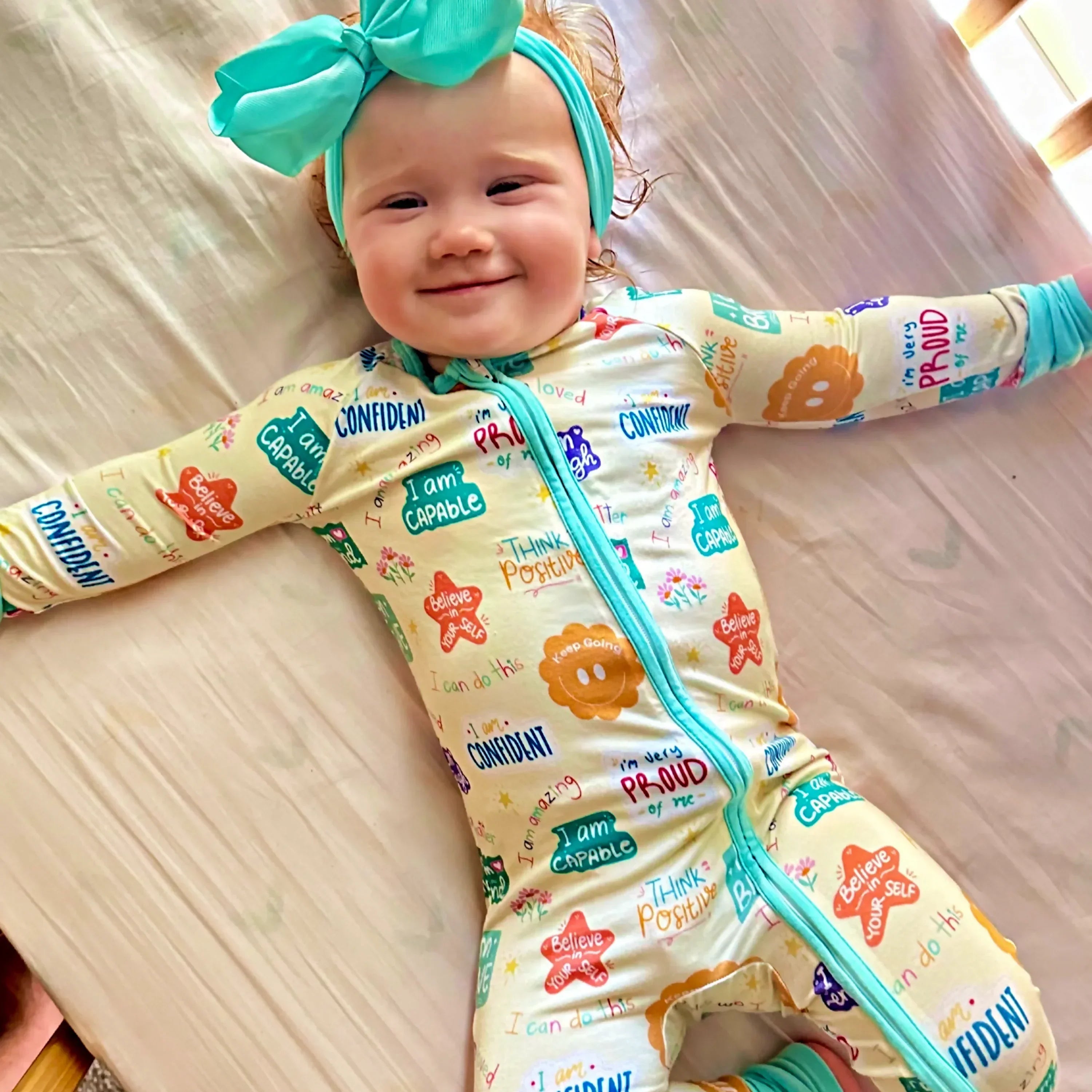 Baby in colorful affirmation pajamas laying on bed with bow