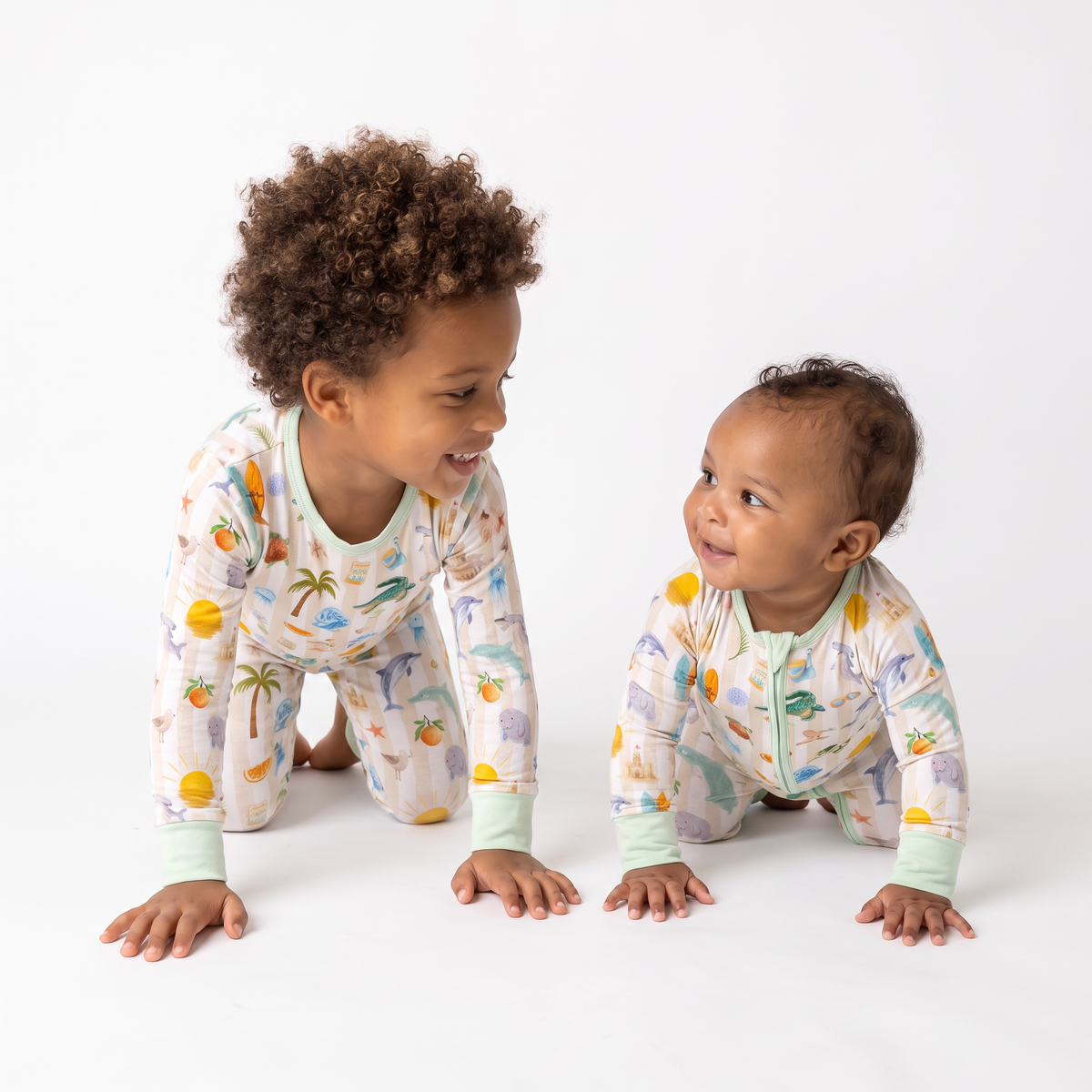 Two children wearing matching printed long-sleeve sleep sacks crawl and smile at each other on a clean white studio background.