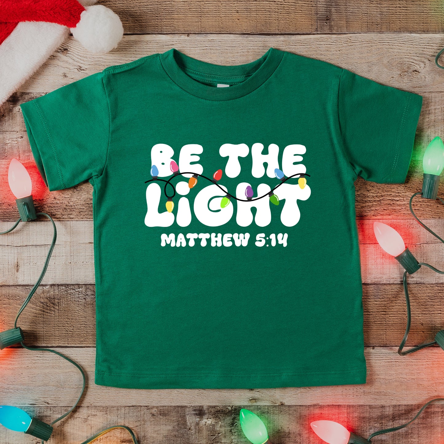 Green t-shirt with 'Be the Light Matthew 5:14' text on a wooden surface with Christmas lights.