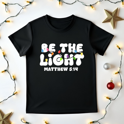 Black t-shirt with 'Be the Light Matthew 5:14' text on a white background with decorative lights and stars.