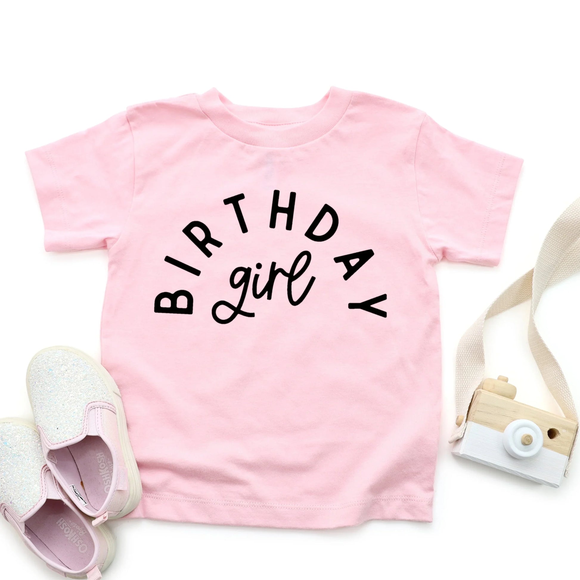 Pink t-shirt with 'Birthday girl' text, white shoes, and a small camera on a white background