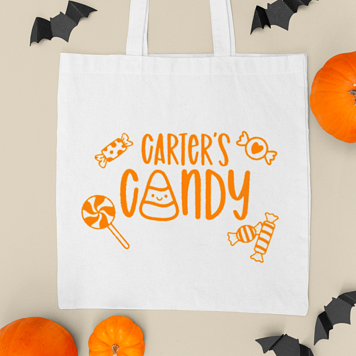 White tote bag with 'Carter's Candy' text and candy graphics on a beige background with Halloween decorations.