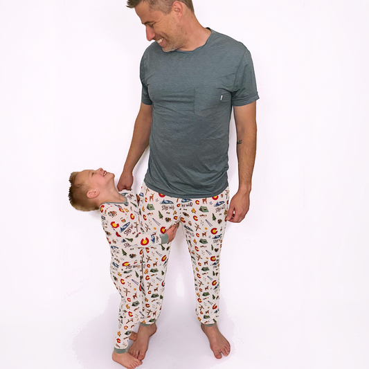A man and a young boy wearing matching pajama sets with a colorful cartoon pattern on a white background. The man wears a solid dark gray t-shirt, while the boy wears a long-sleeve pajama set with an all-over print of red apples, green leaves, and other small illustrations on an off-white base.