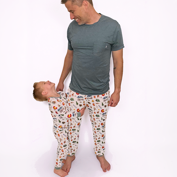 A man and a young boy wearing matching pajama sets with a colorful cartoon pattern on a white background. The man wears a solid dark gray t-shirt, while the boy wears a long-sleeve pajama set with an all-over print of red apples, green leaves, and other small illustrations on an off-white base.