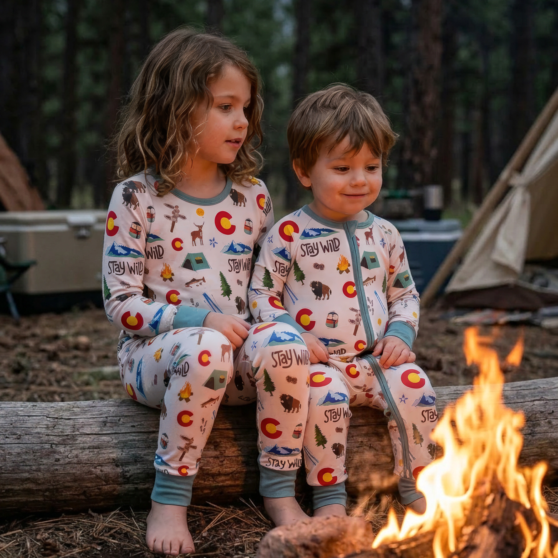 Two children in matching colorado pajamas sitting by a campfire in a forest setting.