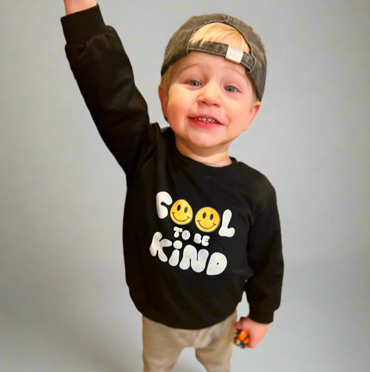 Child wearing a black sweatshirt with smiley faces and text, raising one arm against a plain background