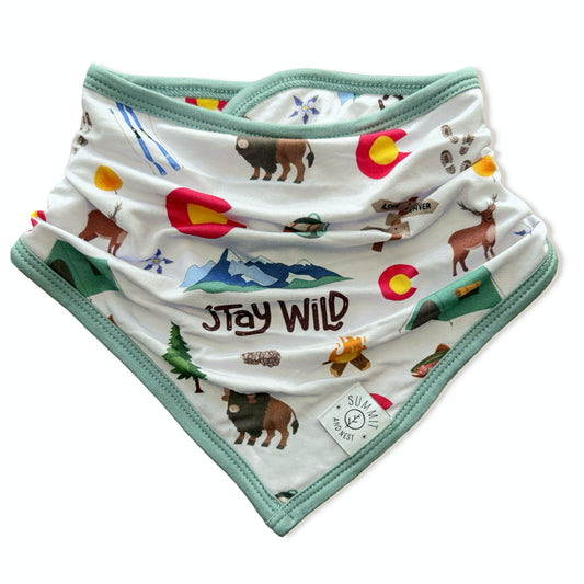 A white baby bandana bib with mint green trim, featuring a colorful outdoor-themed print with the words 'Stay Wild' and various illustrations of animals, mountains, and the Colorado state flag.