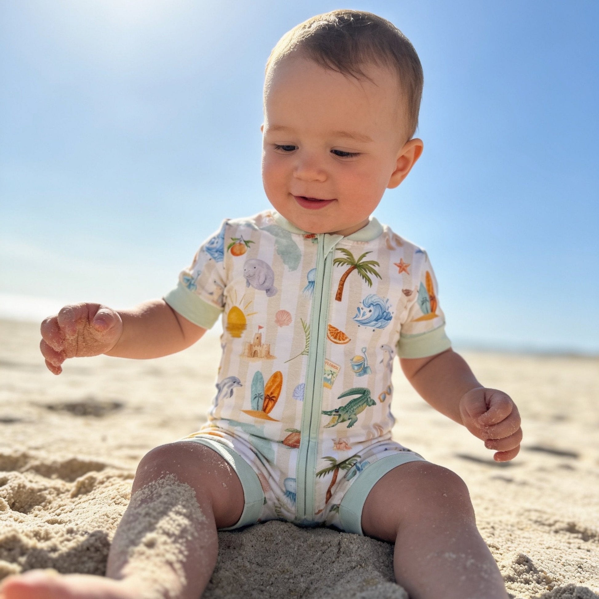 Baby sitting on the sand wearing a Florida romper with sea-themed design.
