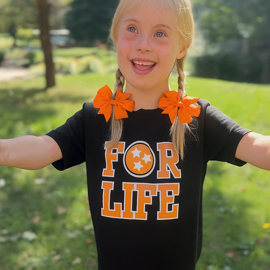 Child wearing a black shirt with 'FOR LIFE' text and orange leggings outdoors.