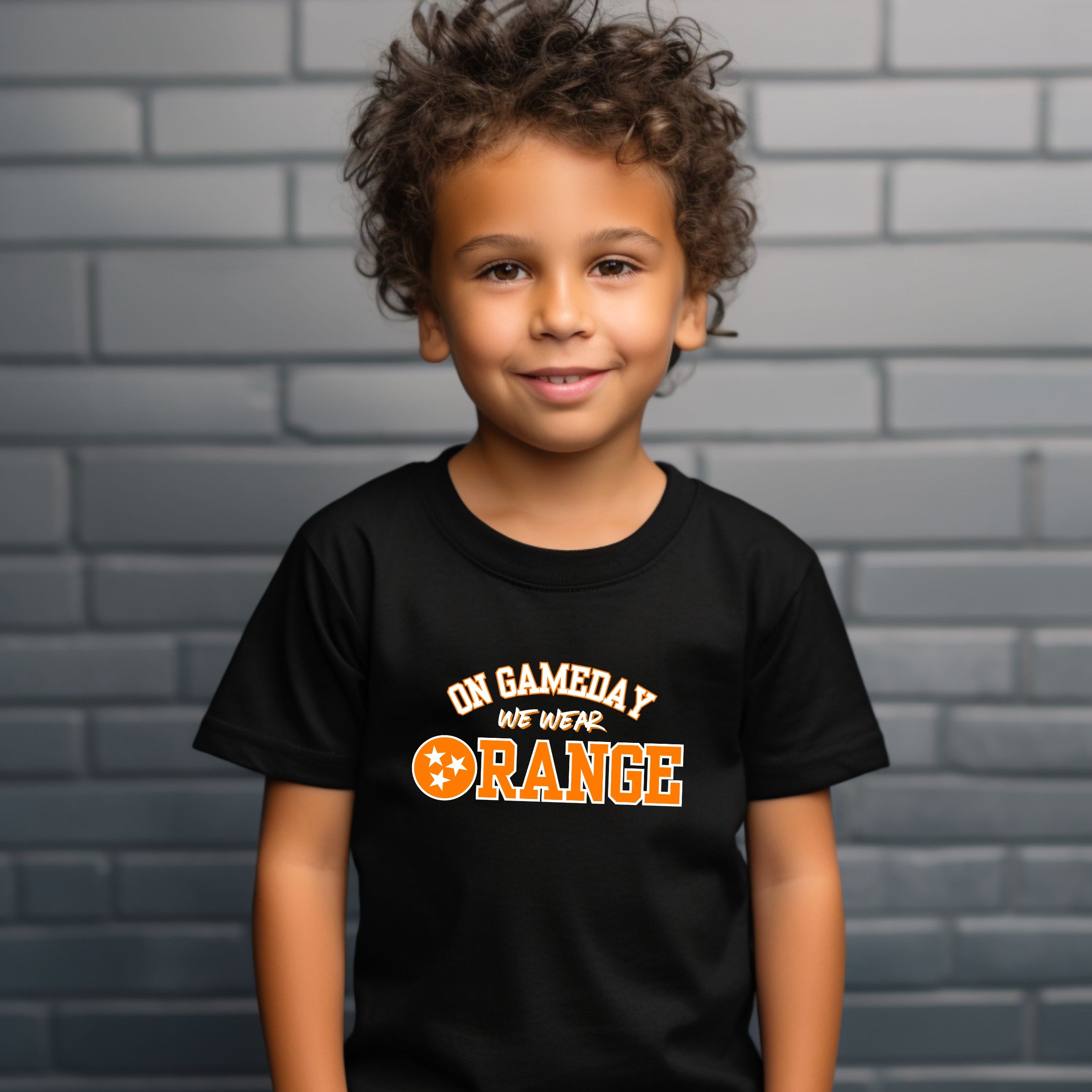 Child wearing a black t-shirt with 'On Gameday We Wear Orange' text, standing against a gray brick wall.