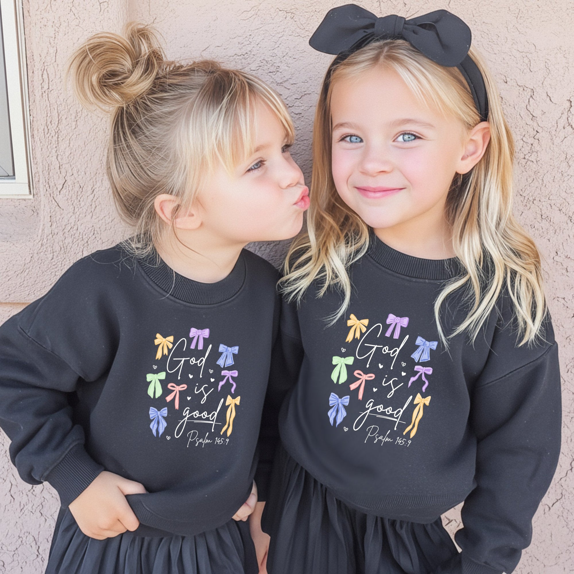 Two young girls wearing matching black God is Good sweatshirts with colorful bow designs, standing against a beige wall