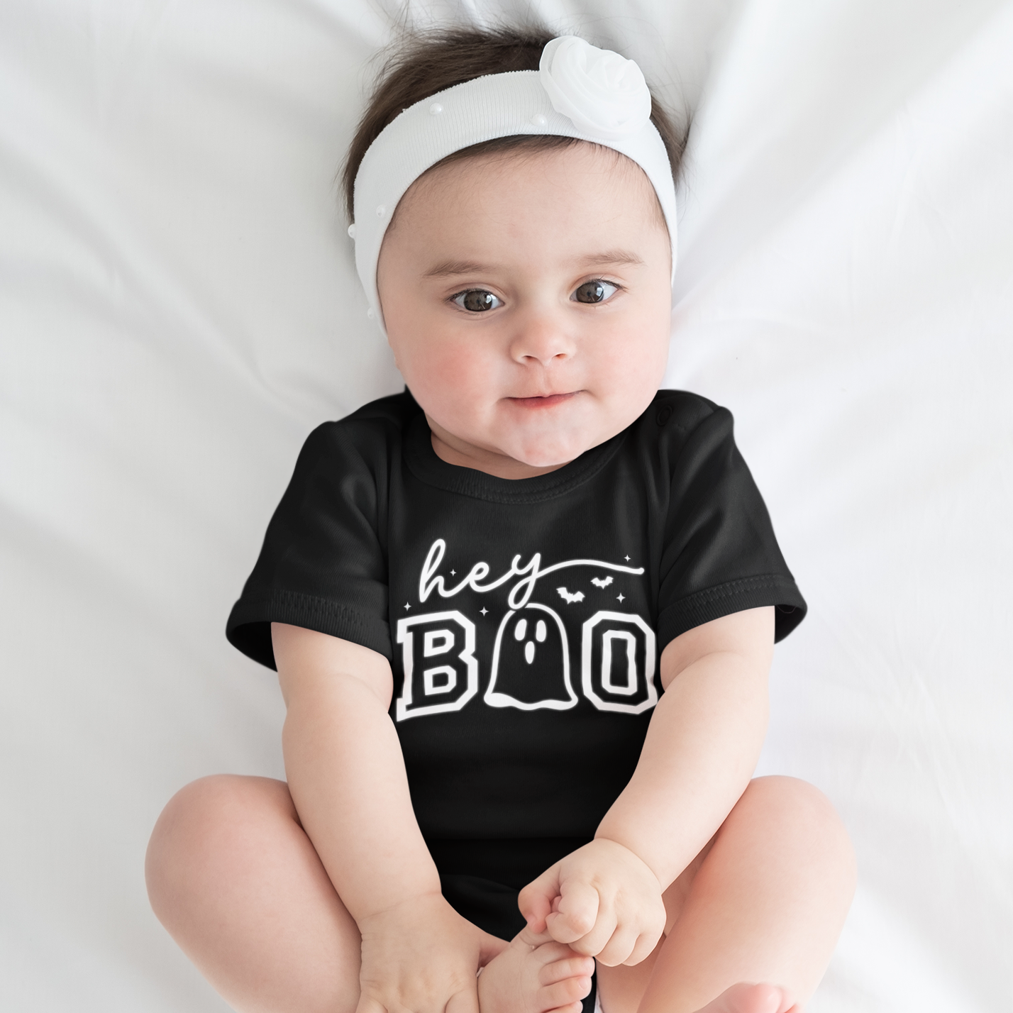 Baby wearing a black onesie with 'hey BOO' text, sitting on a white surface.