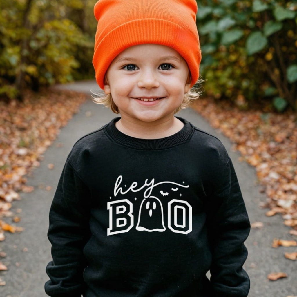 Child wearing a black sweatshirt with 'hey BOO' text, standing outdoors on a path with greenery.