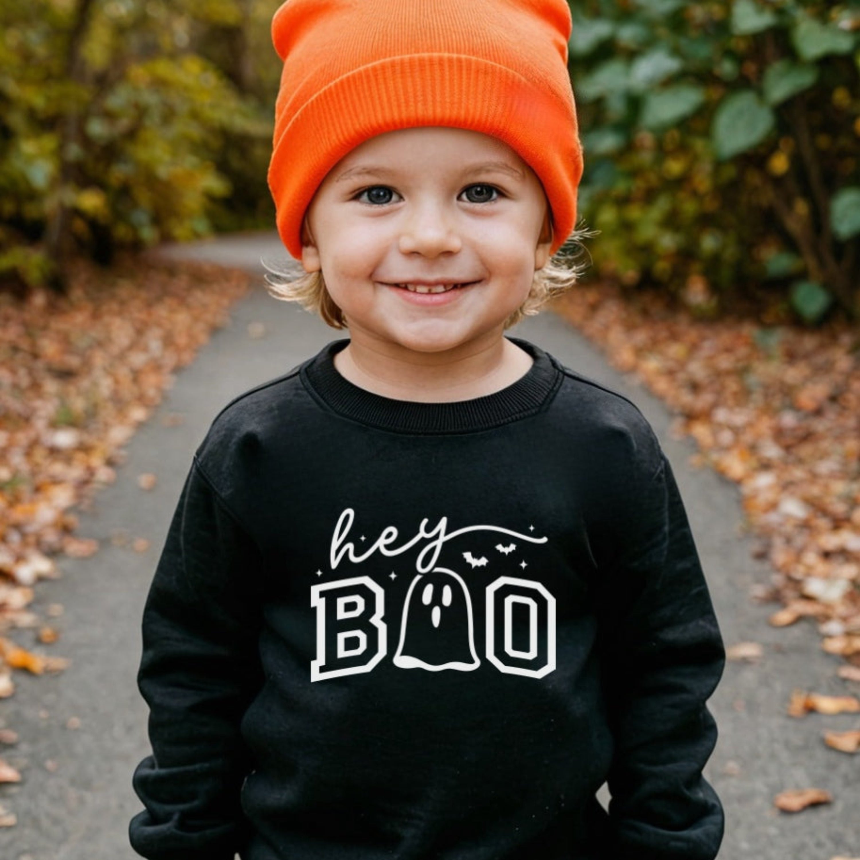 Child wearing a black sweatshirt with 'hey BOO' text, standing outdoors on a path with greenery.