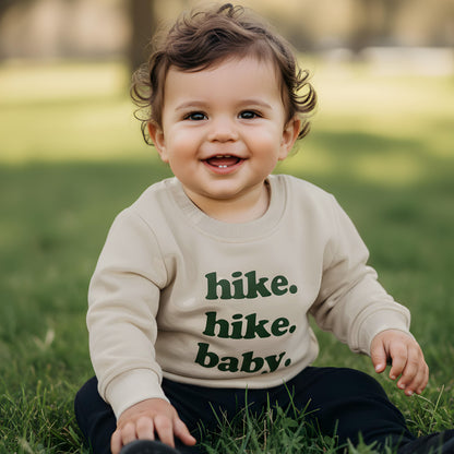 Child wearing a sweater with 'hike. hike. baby.' text, sitting on grass.