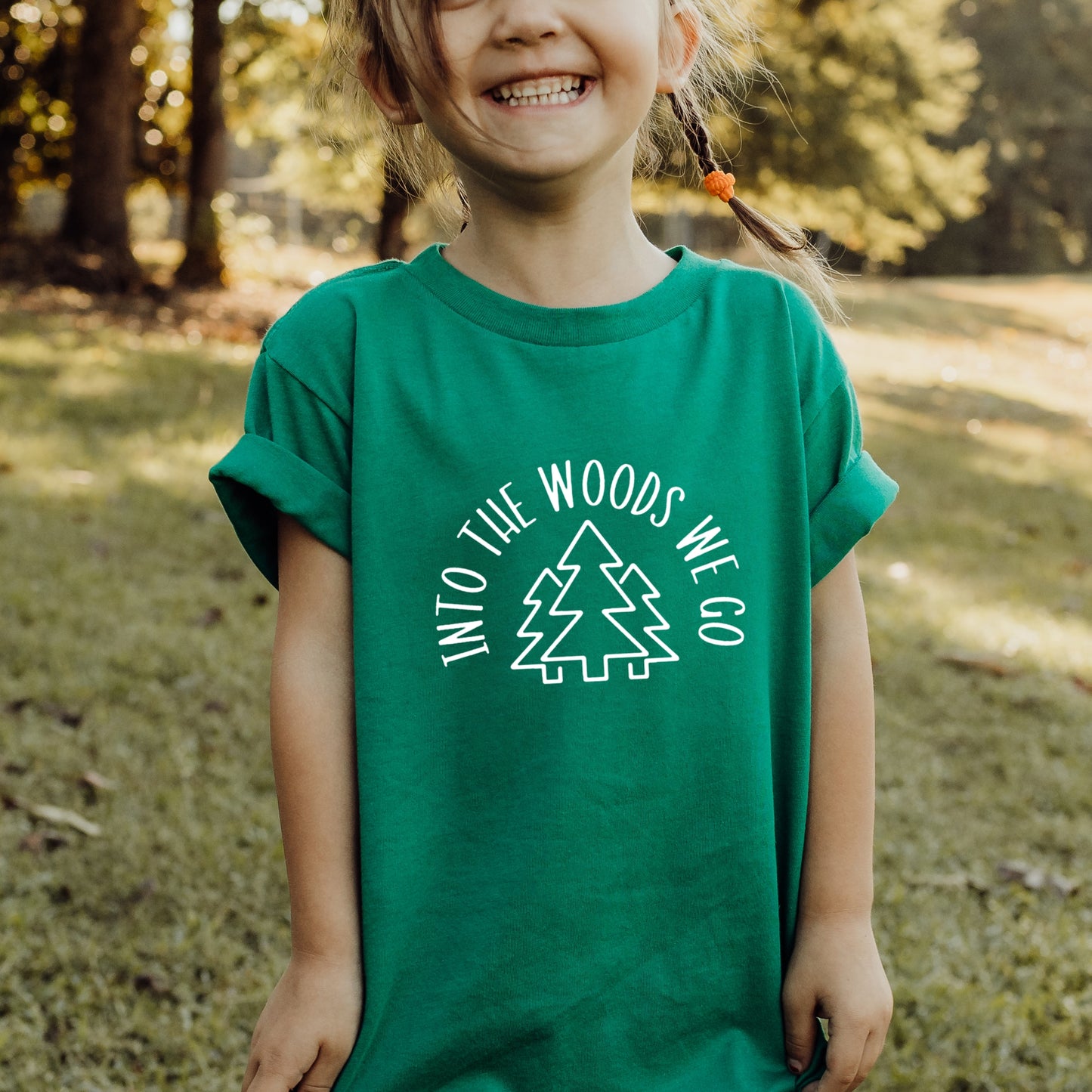 Child wearing a green t-shirt with 'Into the Woods We Go' text and tree graphic outdoors.