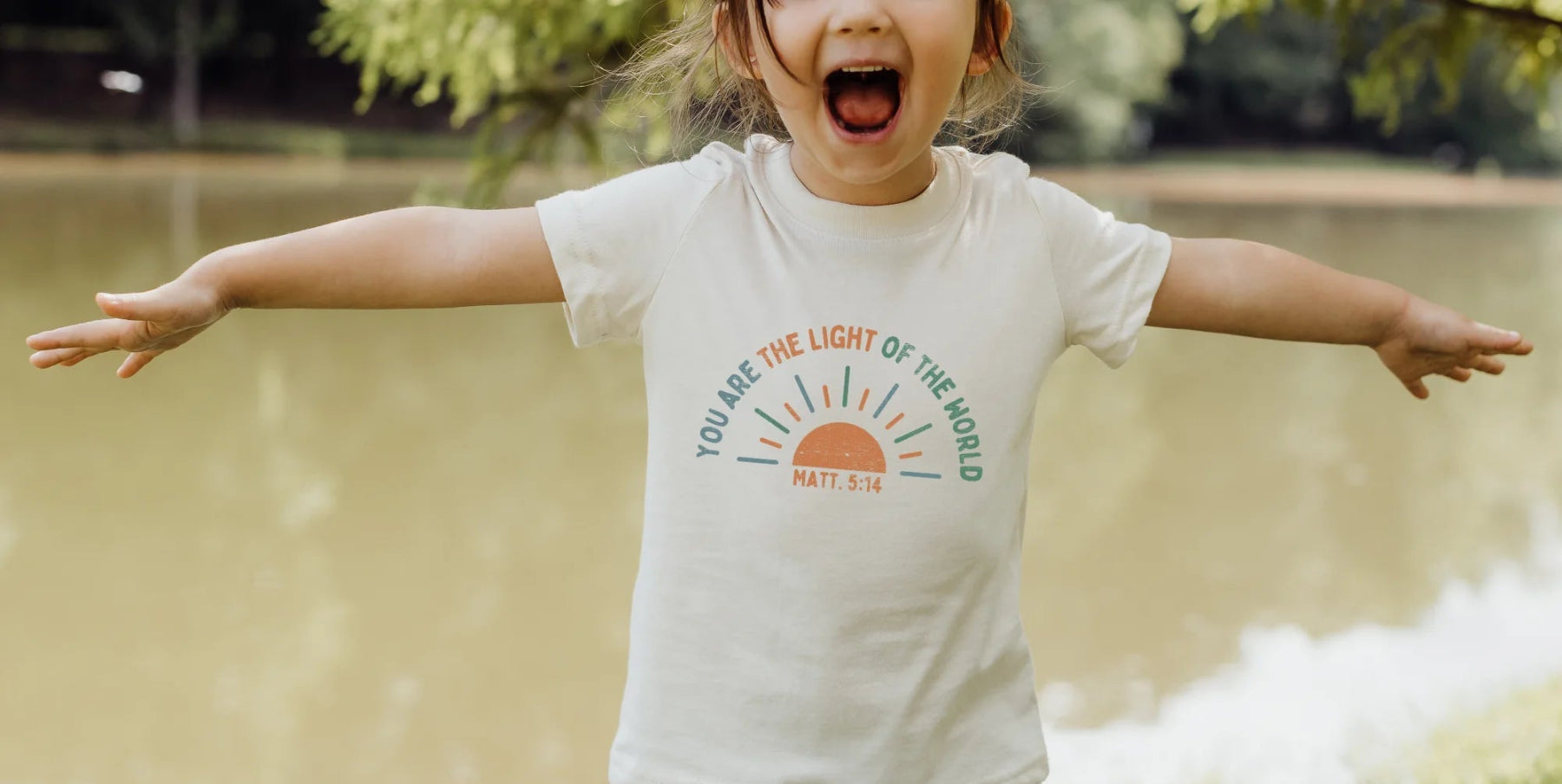 Child wearing a t-shirt with a religious motivational quote outdoors by a lake