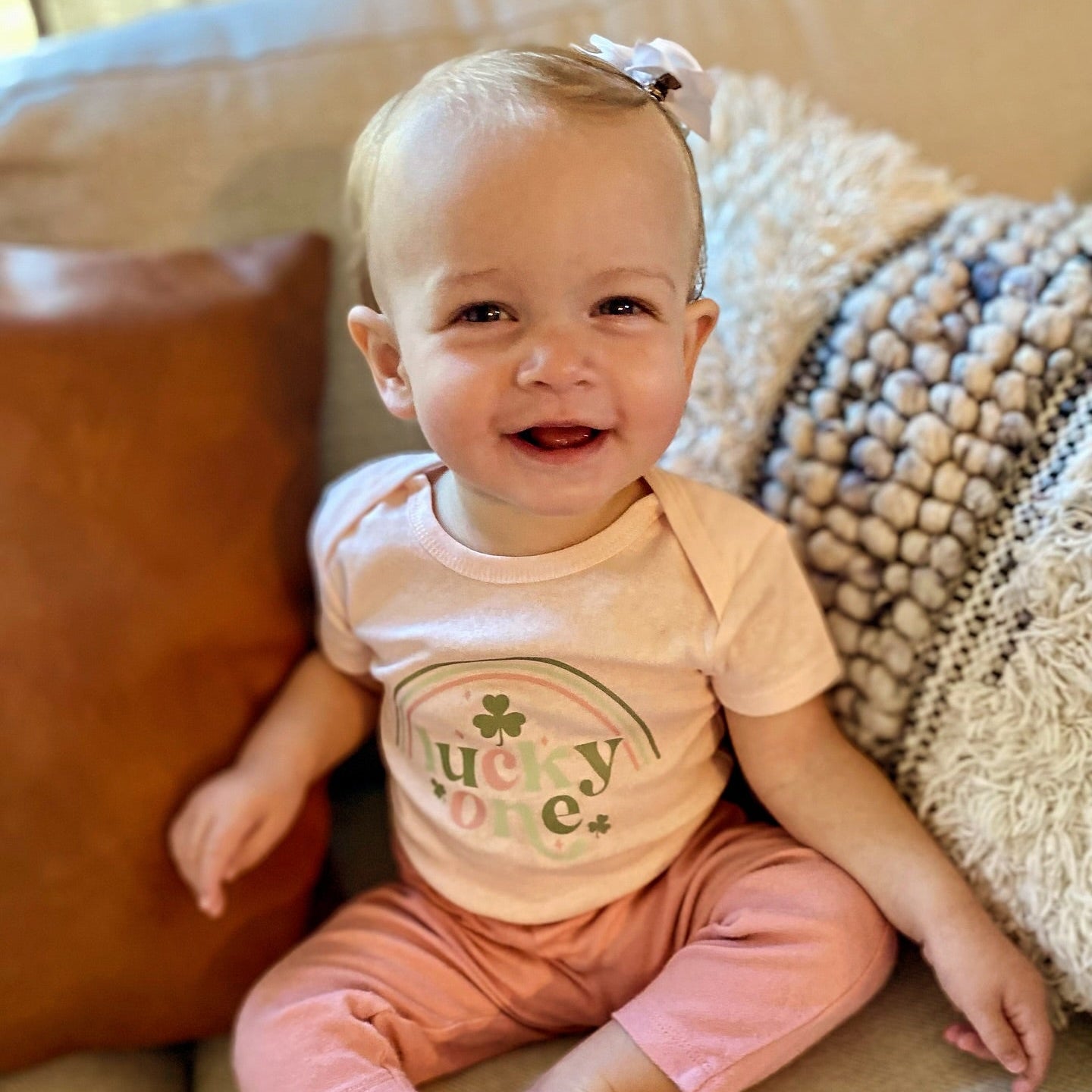 Baby wearing a lucky one pink shirt with a shamrock design sitting on a couch.