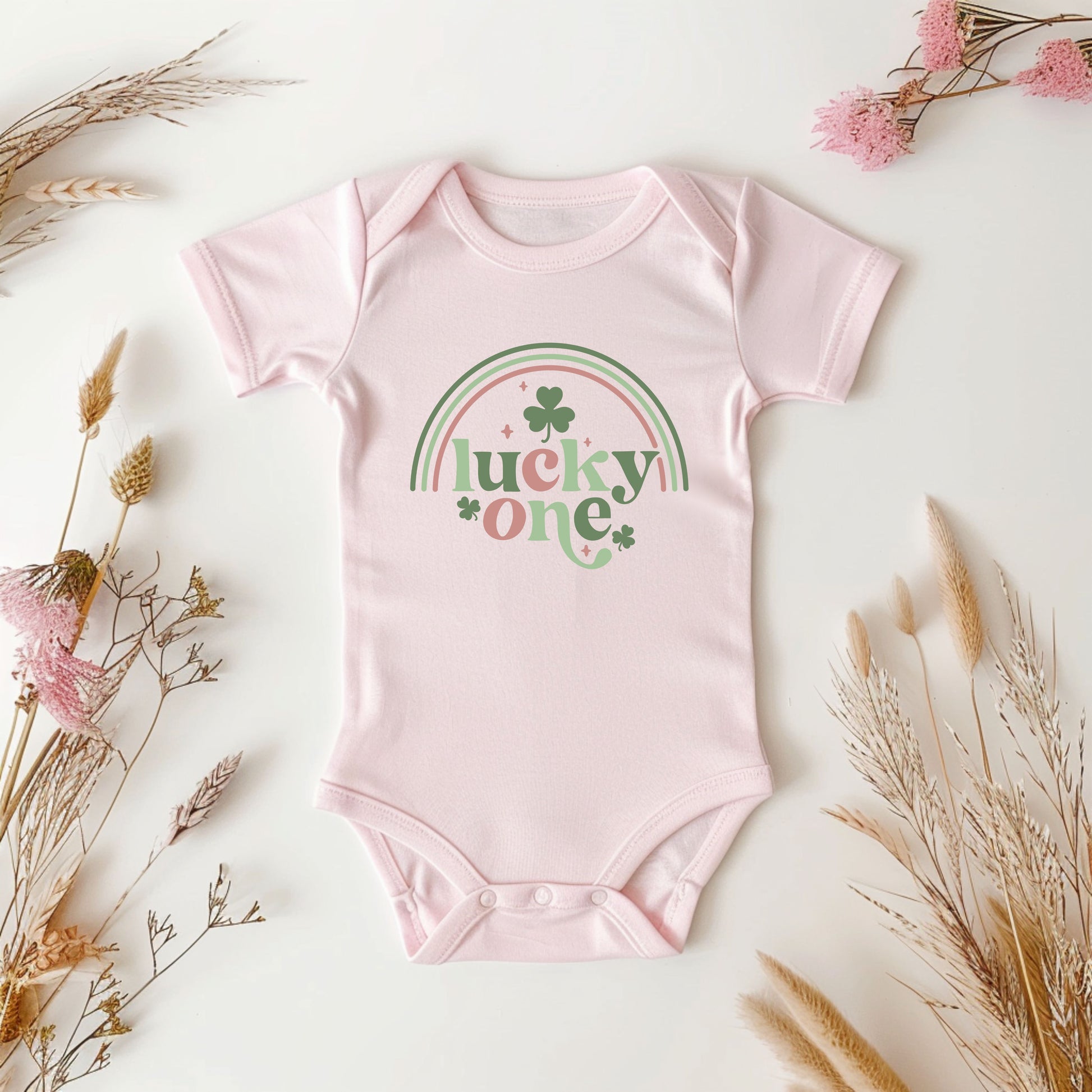 Pink baby onesie with 'lucky one' print on a light background with dried flowers.