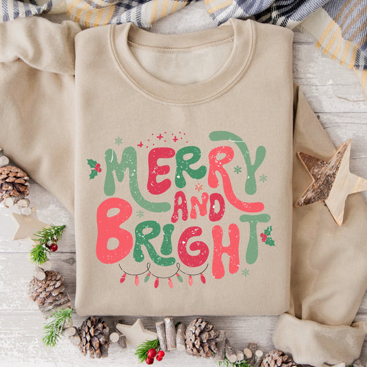 Beige sweatshirt with 'Merry and Bright' text on a wooden surface with Christmas decorations.