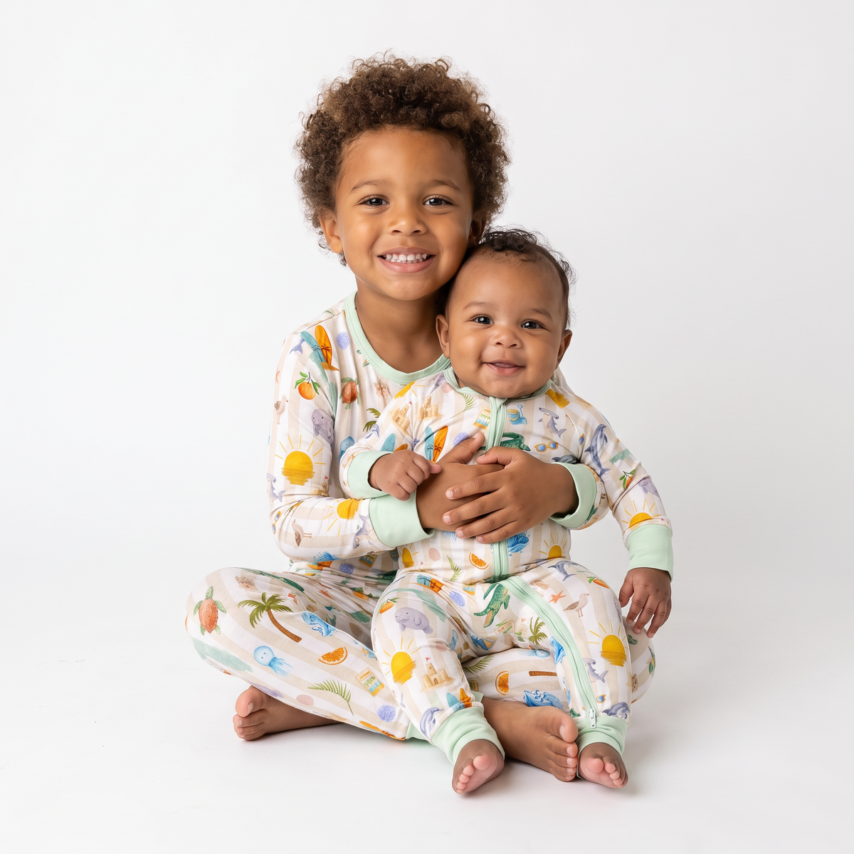 Two children sitting cross-legged on a white studio background, wearing matching one-piece pajamas with a light cream base and a colorful beach-themed print including palm trees, suns, and shells. The older child holds the younger child.
