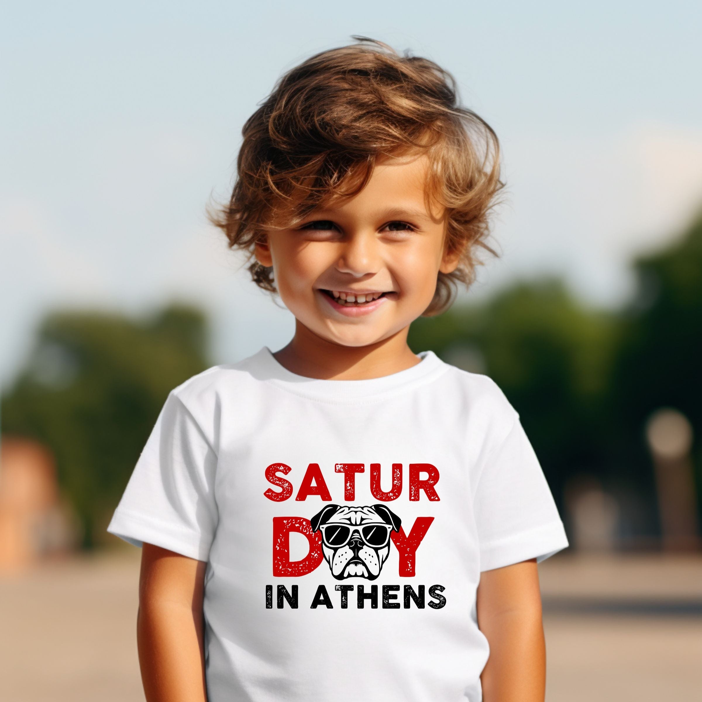 Child wearing a white t-shirt with 'Saturday in Athens' text and graphic, outdoors.