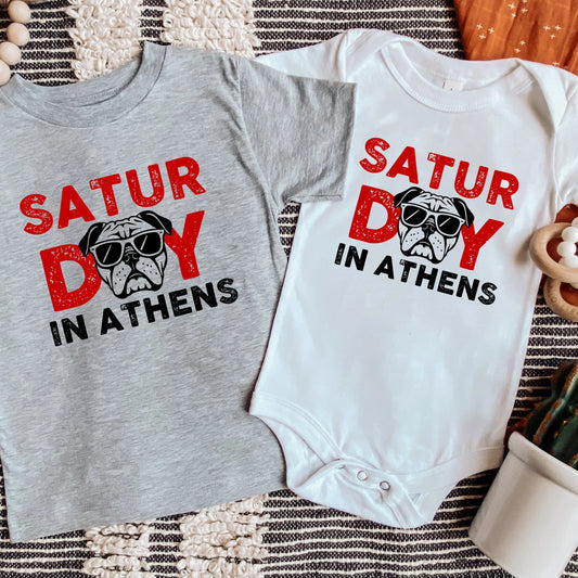 Gray t-shirt and white baby onesie with 'Saturday in Athens' text and pug graphic on a textured surface.
