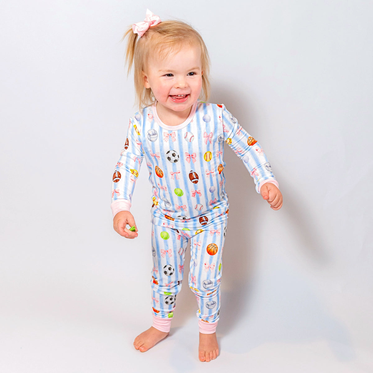 A toddler girl wearing a light blue and white striped long-sleeve pajama set with a colorful sports ball pattern, standing against a light gray studio background.