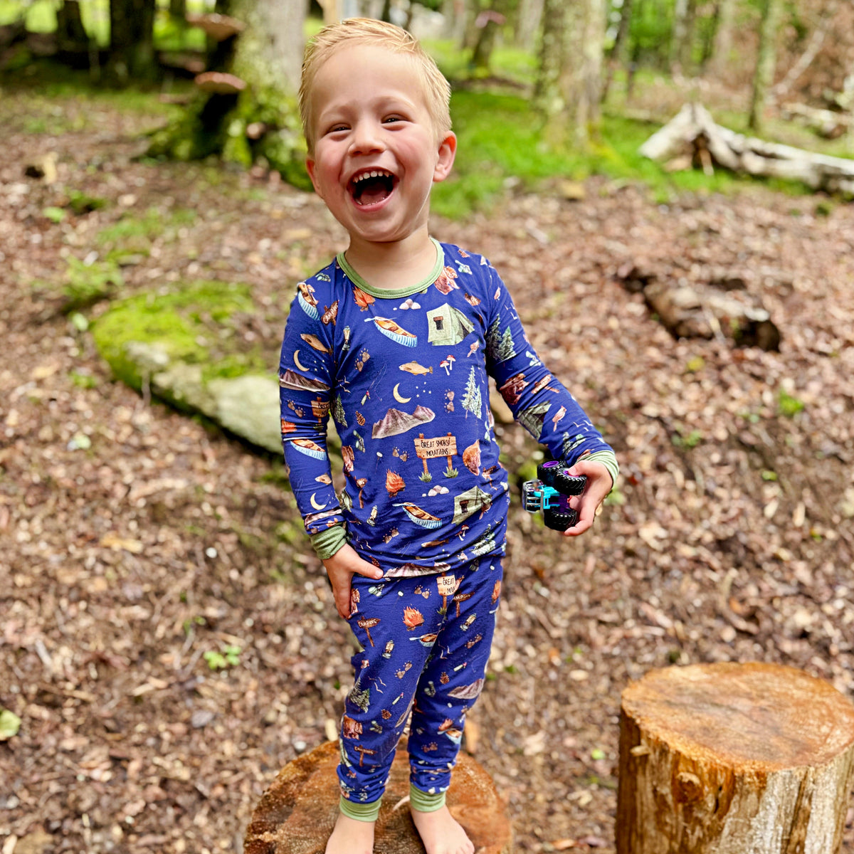 A young boy laughing outdoors in a forest clearing, wearing a blue long-sleeve pajama set with a colorful space-themed print including planets, rockets, stars, and a moon.