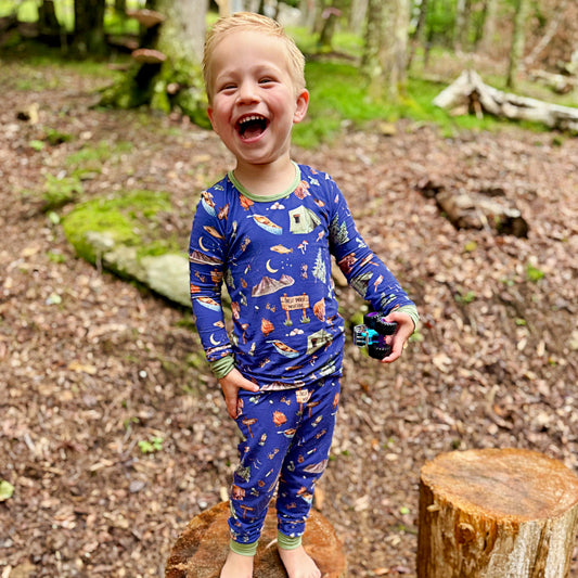A young boy laughing outdoors in a forest clearing, wearing a blue long-sleeve pajama set with a colorful space-themed print including planets, rockets, stars, and a moon.