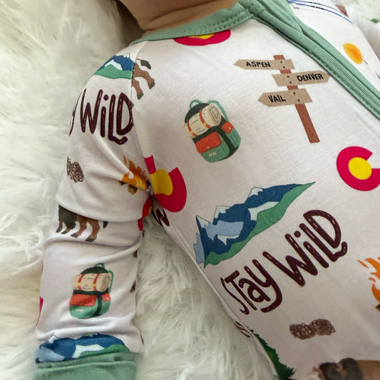 A close-up of a baby wearing a long-sleeve zip-up onesie with a colorful 'Jay Wild' Colorado mountain and trail print, lying on a white faux fur surface.