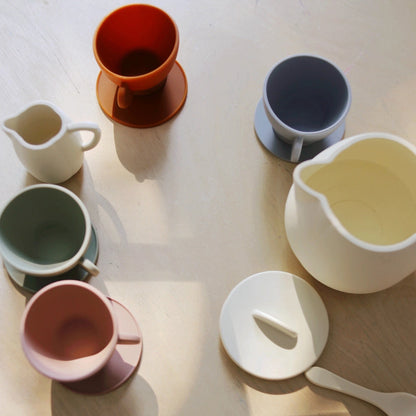 Collection of colorful silicone play  items including cups, a pitcher, and a spoon on a light wooden surface.