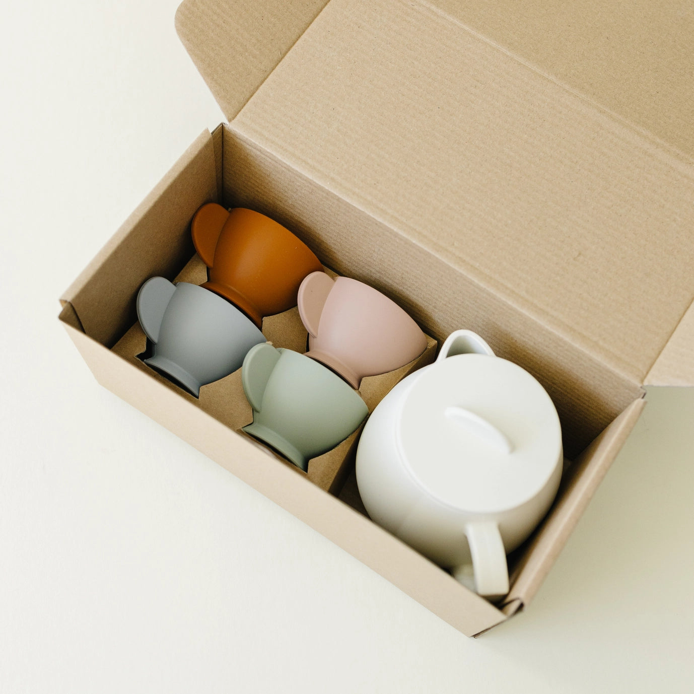 Set of colorful silicone cups and a teapot in a cardboard box on a light background