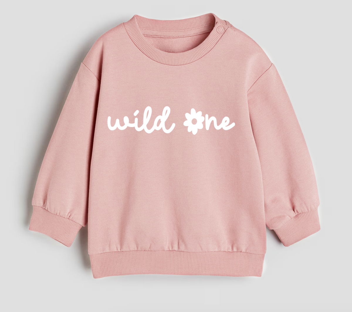 A light pink crewneck sweatshirt with 'wild one' printed in white across the chest, featuring a flower graphic replacing the 'o'.