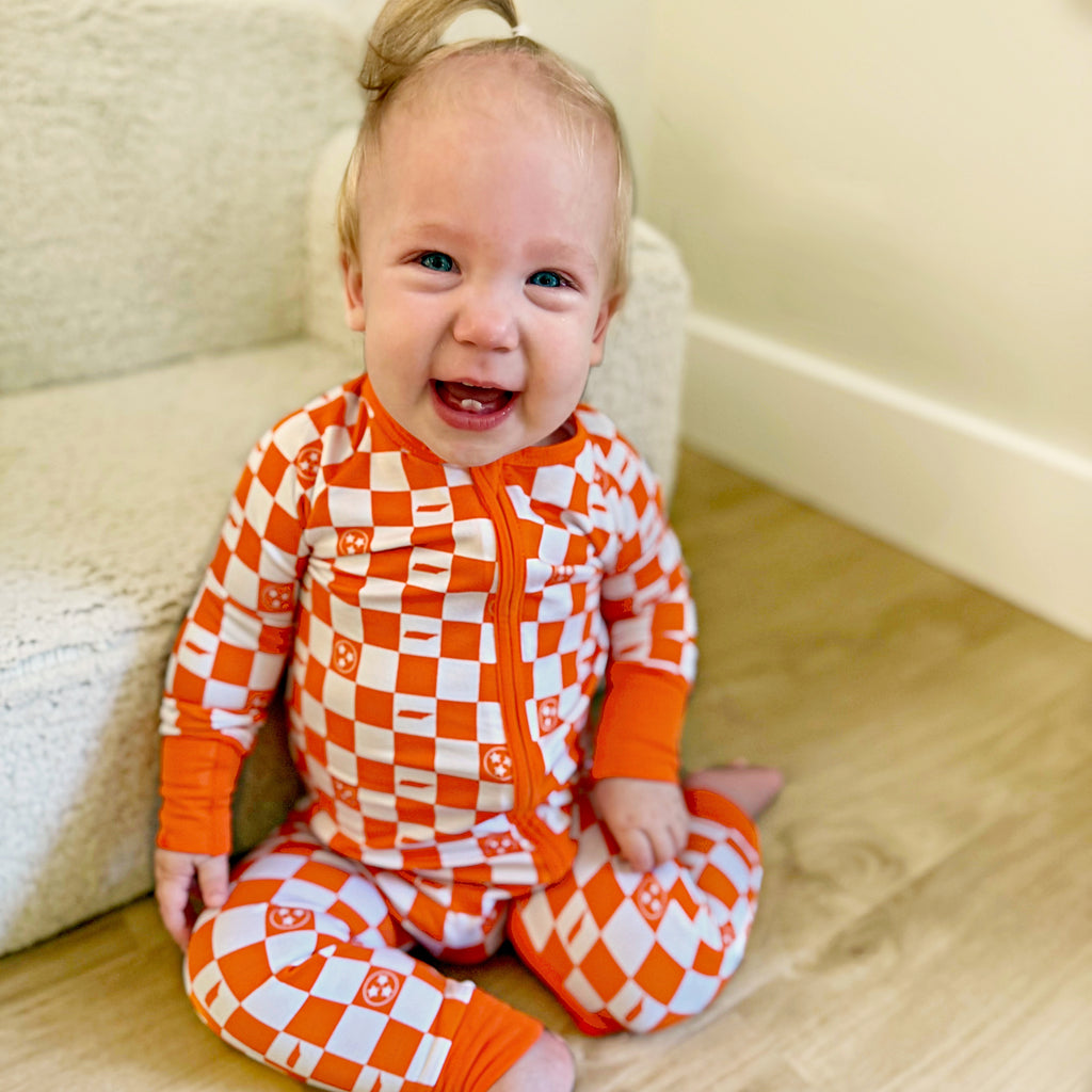 Baby wearing orange and white tennessee checkered pajamas sitting on a wooden floor.